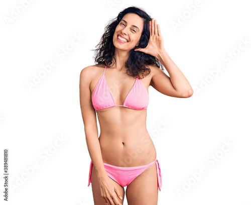 Young beautiful hispanic woman wearing bikini waiving saying hello happy and smiling, friendly welcome gesture