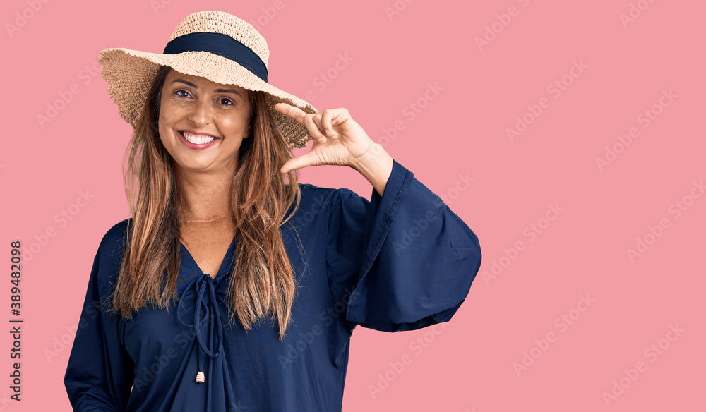 Middle age hispanic woman wearing summer hat smiling and confident gesturing with hand doing small size sign with fingers looking and the camera. measure concept. Middle age hispanic woman wearing summer hat smiling and confident gesturing with hand doing small size sign with fingers looking and the camera. measure concept.