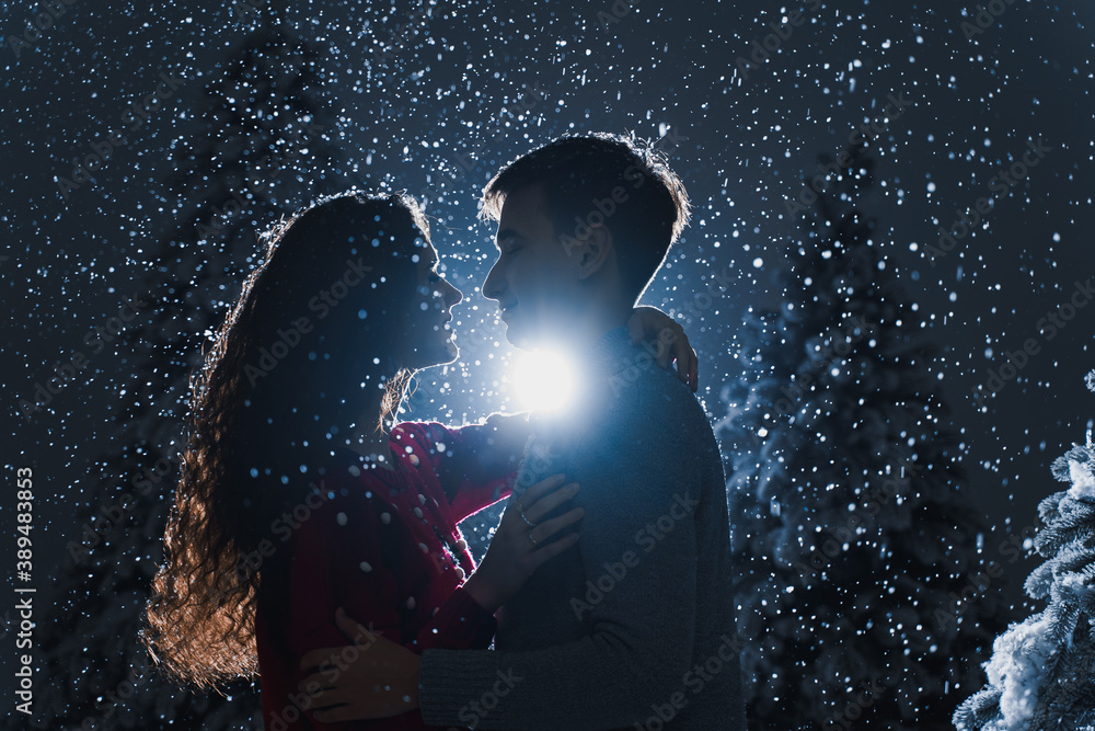 Falling snow and kisses with falling snow and dark blue background. Happy young couple weared white pullovers. hugs and kiss near christmas trees at the eve of new year celebration in winter day Falling snow and kisses with falling snow and dark blue background. Happy young couple weared white pullovers. hugs and kiss near christmas trees at the eve of new year celebration in winter day