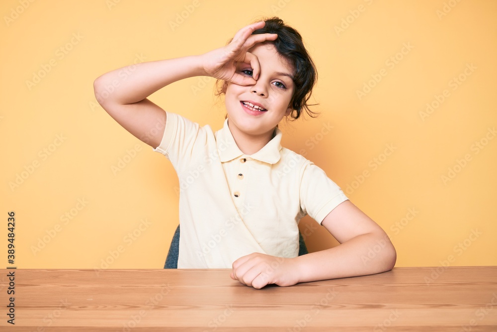 Cute hispanic child wearing casual clothes sitting on the table smiling happy doing ok sign with hand on eye looking through fingers Cute hispanic child wearing casual clothes sitting on the table smiling happy doing ok sign with hand on eye looking through fingers