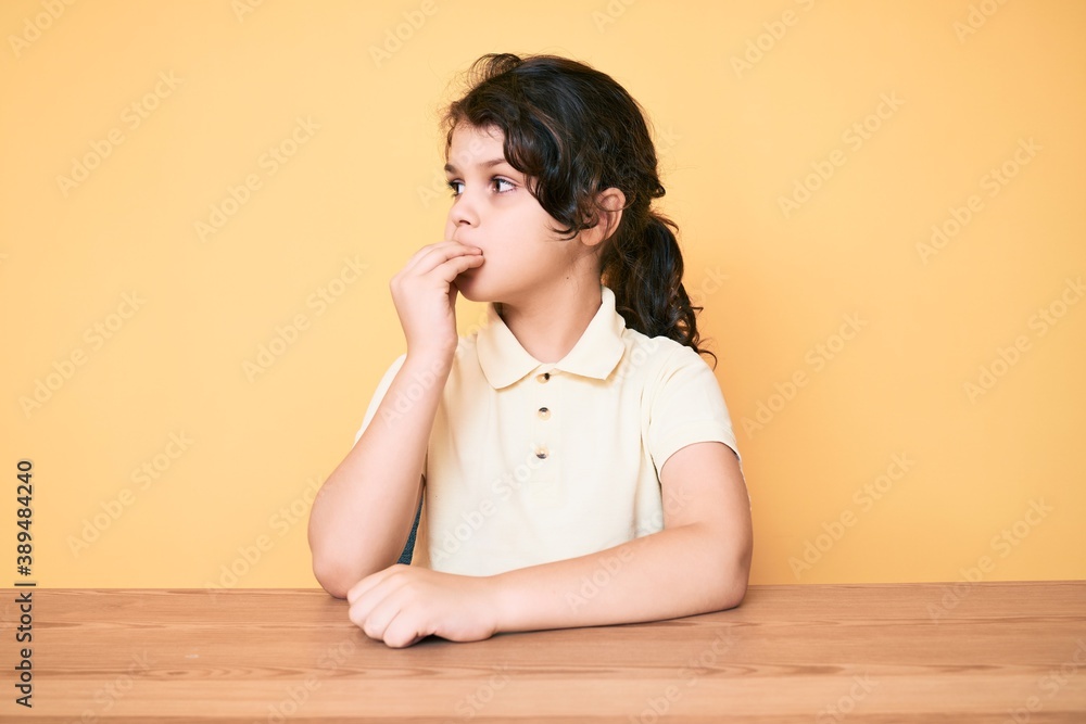 Cute hispanic child wearing casual clothes sitting on the table looking stressed and nervous with hands on mouth biting nails. anxiety problem. Cute hispanic child wearing casual clothes sitting on the table looking stressed and nervous with hands on mouth biting nails. anxiety problem.