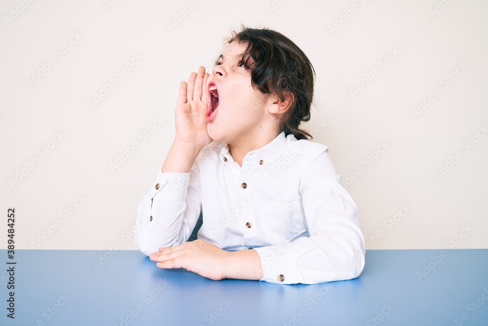 Cute hispanic child wearing casual clothes sitting on the table shouting and screaming loud to side with hand on mouth. communication concept. Cute hispanic child wearing casual clothes sitting on the table shouting and screaming loud to side with hand on mouth. communication concept.