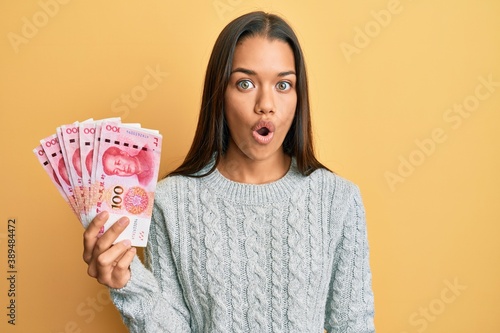 Beautiful hispanic woman holding 100 yuan chinese banknotes scared and amazed with open mouth for surprise, disbelief face