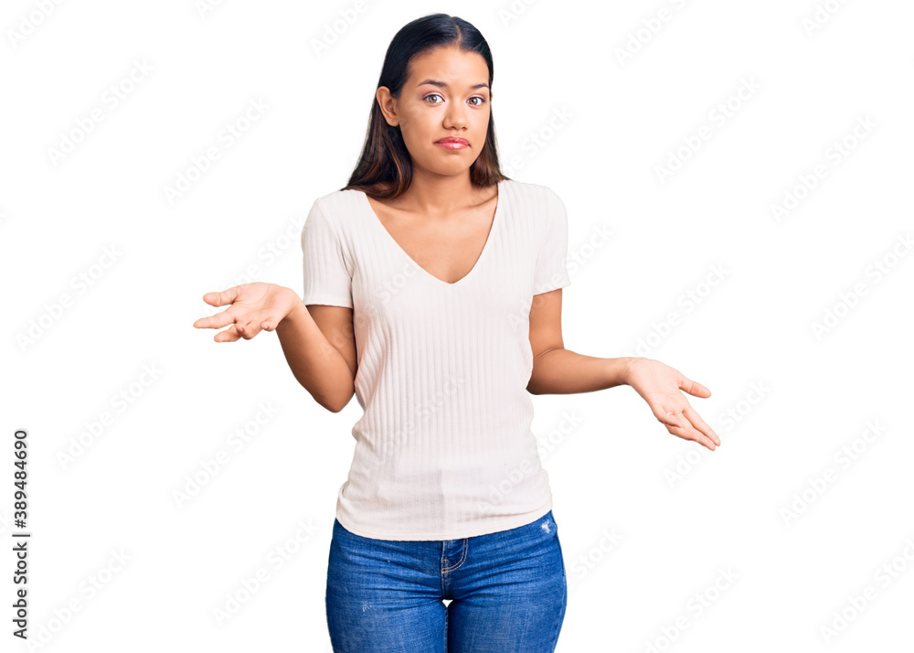 Young beautiful latin girl wearing casual white t shirt clueless and confused expression with arms and hands raised. doubt concept. Young beautiful latin girl wearing casual white t shirt clueless and confused expression with arms and hands raised. doubt concept.