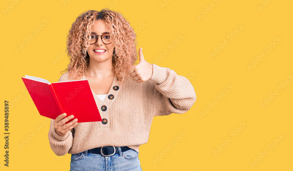 Young blonde woman with curly hair wearing glasses and reading book smiling happy and positive, thumb up doing excellent and approval sign Young blonde woman with curly hair wearing glasses and reading book smiling happy and positive, thumb up doing excellent and approval sign