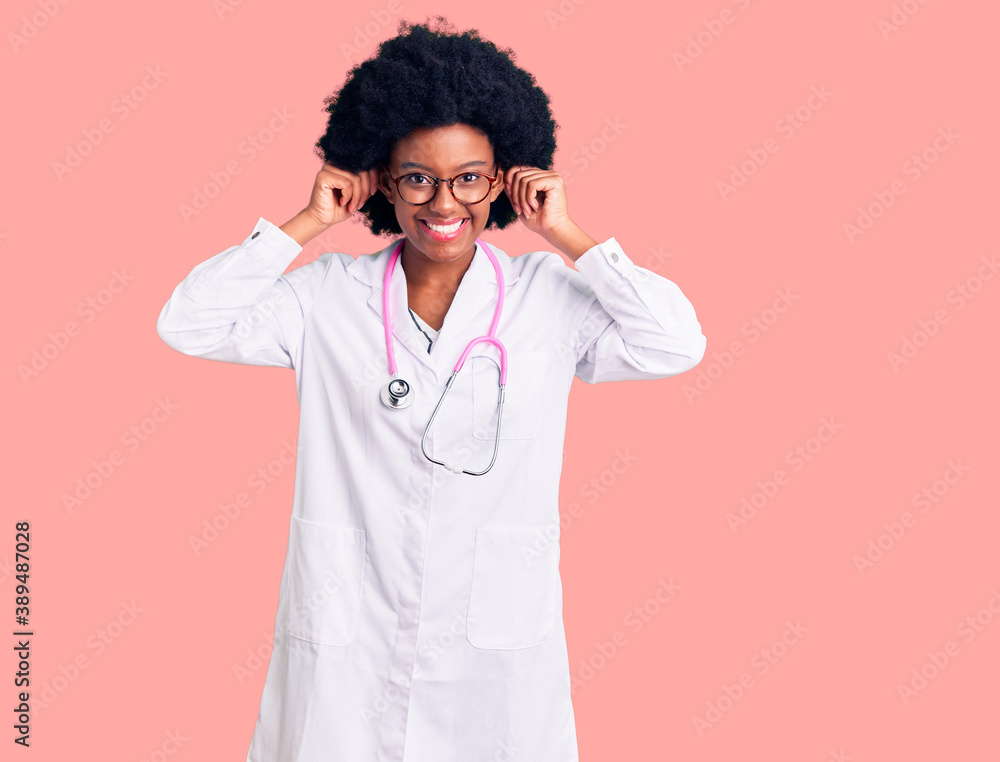 Young african american woman wearing doctor coat and stethoscope smiling pulling ears with fingers, funny gesture. audition problem Young african american woman wearing doctor coat and stethoscope smiling pulling ears with fingers, funny gesture. audition problem