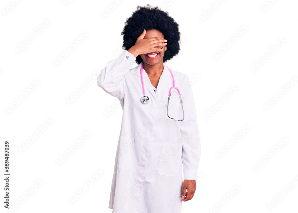 Young african american woman wearing doctor coat and stethoscope smiling and laughing with hand on face covering eyes for surprise. blind concept. Young african american woman wearing doctor coat and stethoscope smiling and laughing with hand on face covering eyes for surprise. blind concept.