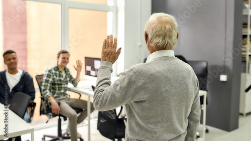 Back view of aged man, senior intern waving, greeting young colleagues at his new job in the modern office