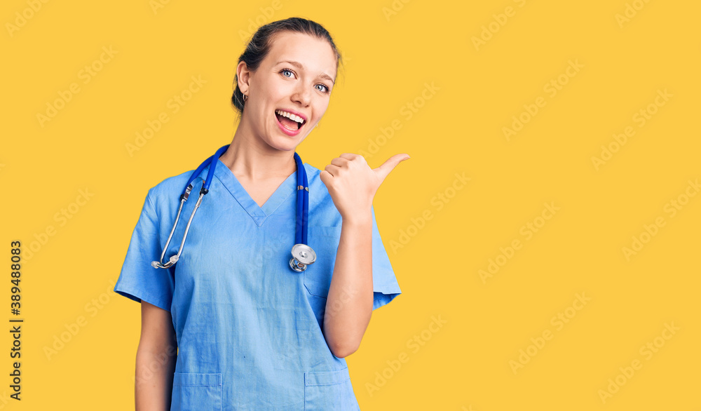 Young beautiful blonde woman wearing doctor uniform and stethoscope smiling with happy face looking and pointing to the side with thumb up. Young beautiful blonde woman wearing doctor uniform and stethoscope smiling with happy face looking and pointing to the side with thumb up.