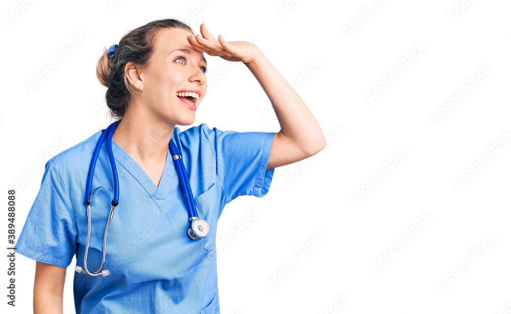 Young beautiful blonde woman wearing doctor uniform and stethoscope very happy and smiling looking far away with hand over head. searching concept. Young beautiful blonde woman wearing doctor uniform and stethoscope very happy and smiling looking far away with hand over head. searching concept.