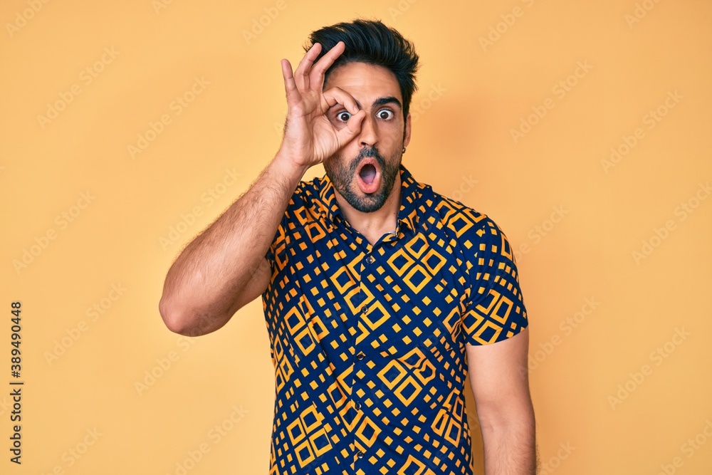 Handsome hispanic man with beard wearing 80s disco style shirt doing ok gesture shocked with surprised face, eye looking through fingers. unbelieving expression. Handsome hispanic man with beard wearing 80s disco style shirt doing ok gesture shocked with surprised face, eye looking through fingers. unbelieving expression.