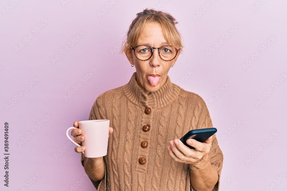 Middle age blonde woman using smartphone and drinking a cup of coffee sticking tongue out happy with funny expression. Middle age blonde woman using smartphone and drinking a cup of coffee sticking tongue out happy with funny expression.