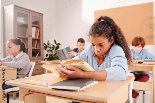 Clever cute schoolgirl reading book and making notes by desk in classroom