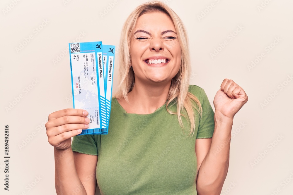 Beautiful blonde tourist woman on vacation holding airline boarding pass over white background screaming proud, celebrating victory and success very excited with raised arm Beautiful blonde tourist woman on vacation holding airline boarding pass over white background screaming proud, celebrating victory and success very excited with raised arm