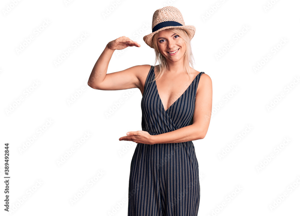 Young beautiful blonde woman wearing summer hat and dress gesturing with hands showing big and large size sign, measure symbol. smiling looking at the camera. measuring concept. Young beautiful blonde woman wearing summer hat and dress gesturing with hands showing big and large size sign, measure symbol. smiling looking at the camera. measuring concept.