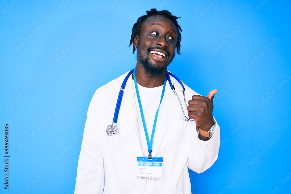 Young african american man with braids wearing doctor stethoscope and id pass smiling with happy face looking and pointing to the side with thumb up. Young african american man with braids wearing doctor stethoscope and id pass smiling with happy face looking and pointing to the side with thumb up.