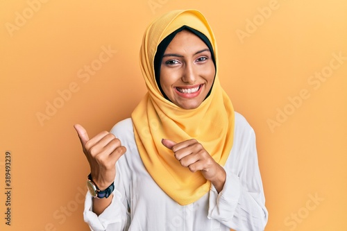 Young brunette arab woman wearing traditional islamic hijab scarf pointing to the back behind with hand and thumbs up, smiling confident © Krakenimages.com