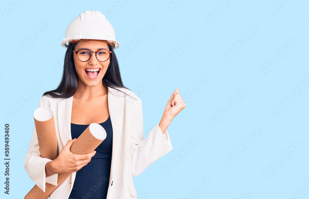 Young beautiful latin girl wearing architect hardhat holding blueprints screaming proud, celebrating victory and success very excited with raised arms Young beautiful latin girl wearing architect hardhat holding blueprints screaming proud, celebrating victory and success very excited with raised arms
