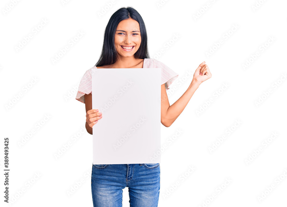 Young beautiful latin girl holding blank empty banner screaming proud, celebrating victory and success very excited with raised arms Young beautiful latin girl holding blank empty banner screaming proud, celebrating victory and success very excited with raised arms