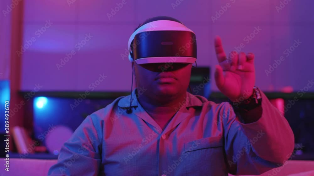 Serious mixed race young man wearing virtual reality headset sitting on sofa at home neon light. Fun game. VR glasses Social network robot. Slow motion Serious mixed race young man wearing virtual reality headset sitting on sofa at home neon light. Fun game. VR glasses Social network robot. Slow motion