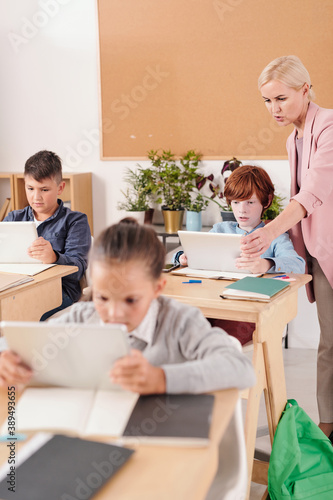 Young confident teacher in elegant suit consulting cute schoolboy with tablet