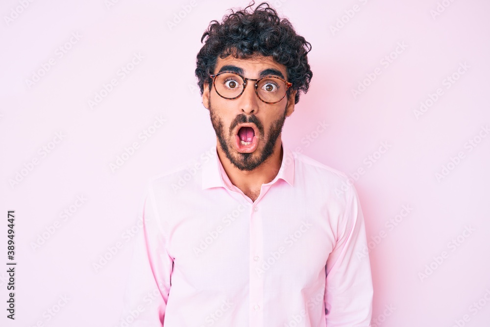 Handsome young man with curly hair and bear wearing business shirt and glasses scared and amazed with open mouth for surprise, disbelief face Handsome young man with curly hair and bear wearing business shirt and glasses scared and amazed with open mouth for surprise, disbelief face