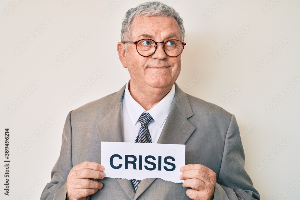 Senior grey-haired man wearing business suit holding crisis paper smiling looking to the side and staring away thinking. Senior grey-haired man wearing business suit holding crisis paper smiling looking to the side and staring away thinking.
