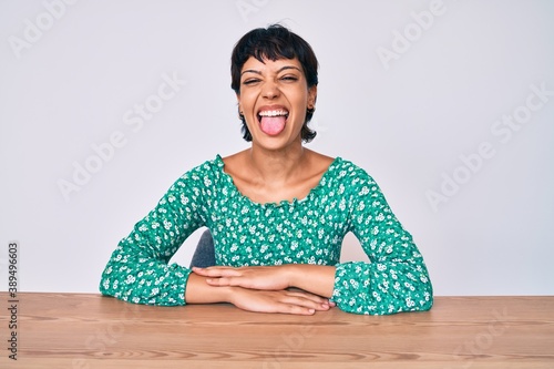 Beautiful brunettte woman wearing casual clothes sitting on the table sticking tongue out happy with funny expression. emotion concept.