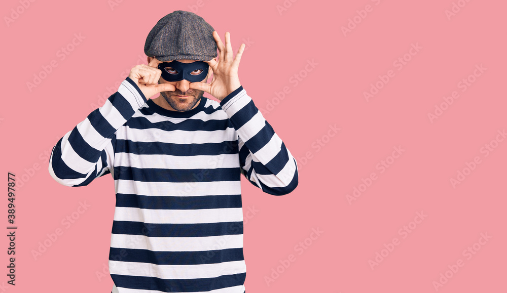 Young handsome man wearing burglar mask trying to open eyes with fingers, sleepy and tired for morning fatigue Young handsome man wearing burglar mask trying to open eyes with fingers, sleepy and tired for morning fatigue