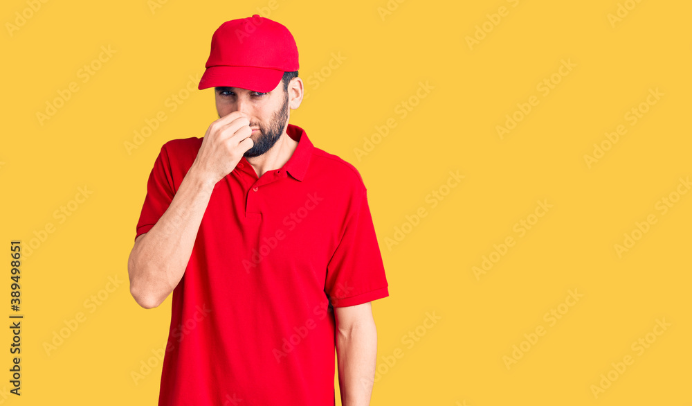 Young handsome man with beard wearing delivery uniform smelling something stinky and disgusting, intolerable smell, holding breath with fingers on nose. bad smell
