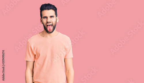 Young handsome man with beard wearing casual t-shirt sticking tongue out happy with funny expression. emotion concept.