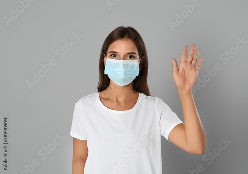 Woman in protective mask showing hello gesture on grey background. Keeping social distance during coronavirus pandemic