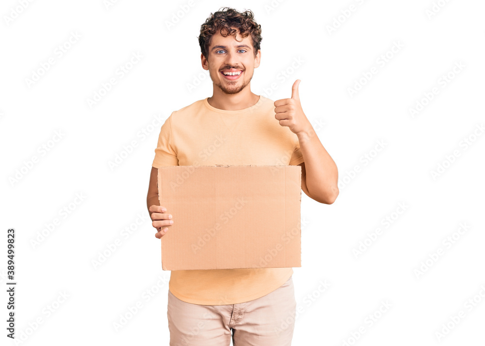 Young handsome man with curly hair holding blank cardboard empty banner smiling happy and positive, thumb up doing excellent and approval sign Young handsome man with curly hair holding blank cardboard empty banner smiling happy and positive, thumb up doing excellent and approval sign