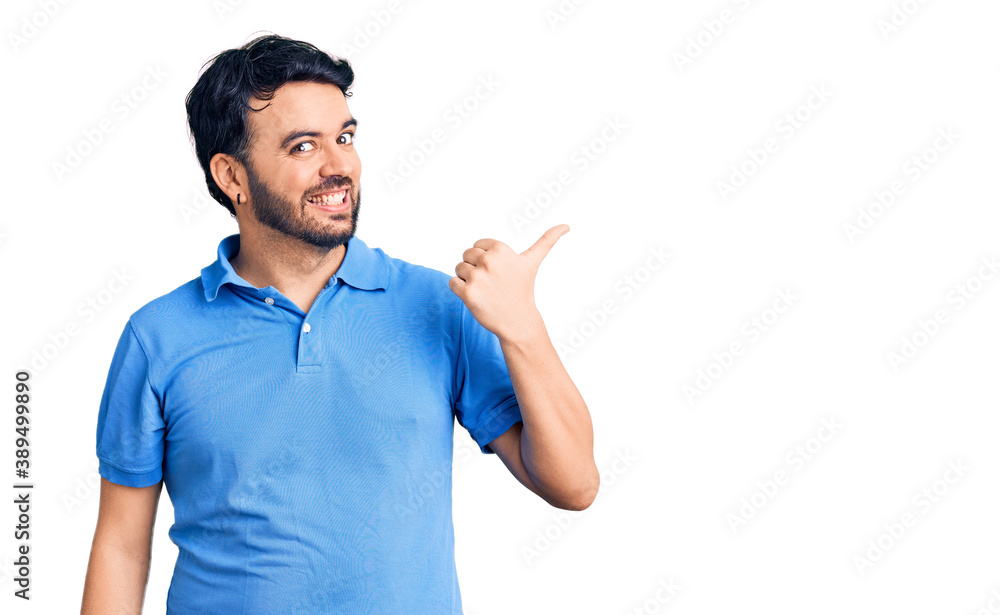 Young hispanic man wearing casual clothes smiling with happy face looking and pointing to the side with thumb up. Young hispanic man wearing casual clothes smiling with happy face looking and pointing to the side with thumb up.