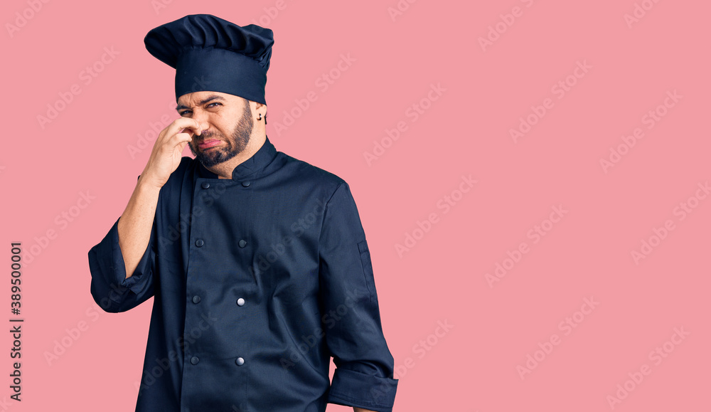 Young hispanic man wearing cooker uniform smelling something stinky and disgusting, intolerable smell, holding breath with fingers on nose. bad smell Young hispanic man wearing cooker uniform smelling something stinky and disgusting, intolerable smell, holding breath with fingers on nose. bad smell