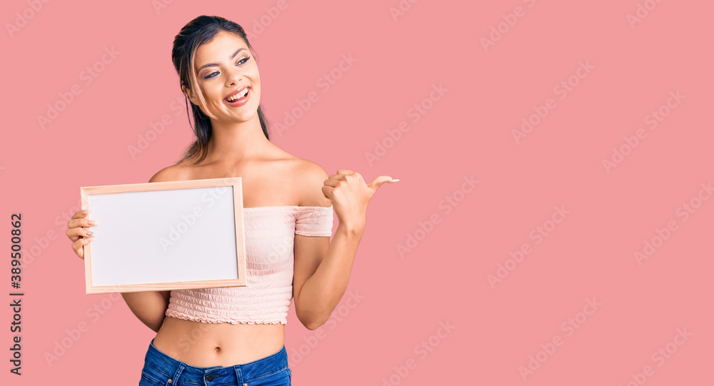 Young beautiful woman holding empty white chalkboard pointing thumb up to the side smiling happy with open mouth Young beautiful woman holding empty white chalkboard pointing thumb up to the side smiling happy with open mouth