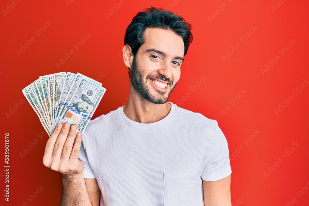 Young hispanic man holding dollars looking positive and happy standing and smiling with a confident smile showing teeth Young hispanic man holding dollars looking positive and happy standing and smiling with a confident smile showing teeth