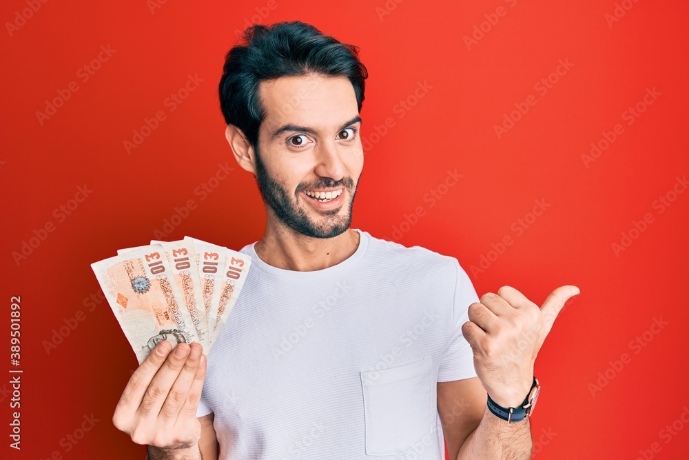 Young hispanic man holding 10 united kingdom pounds banknotes pointing thumb up to the side smiling happy with open mouth Young hispanic man holding 10 united kingdom pounds banknotes pointing thumb up to the side smiling happy with open mouth