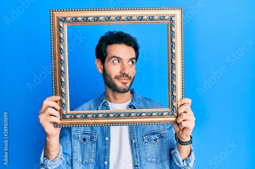 Young hispanic man holding empty frame smiling looking to the side and staring away thinking. photo