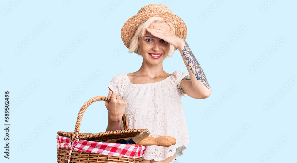 Young blonde woman with tattoo wearing summer hat and holding picnic wicker basket with bread stressed and frustrated with hand on head, surprised and angry face