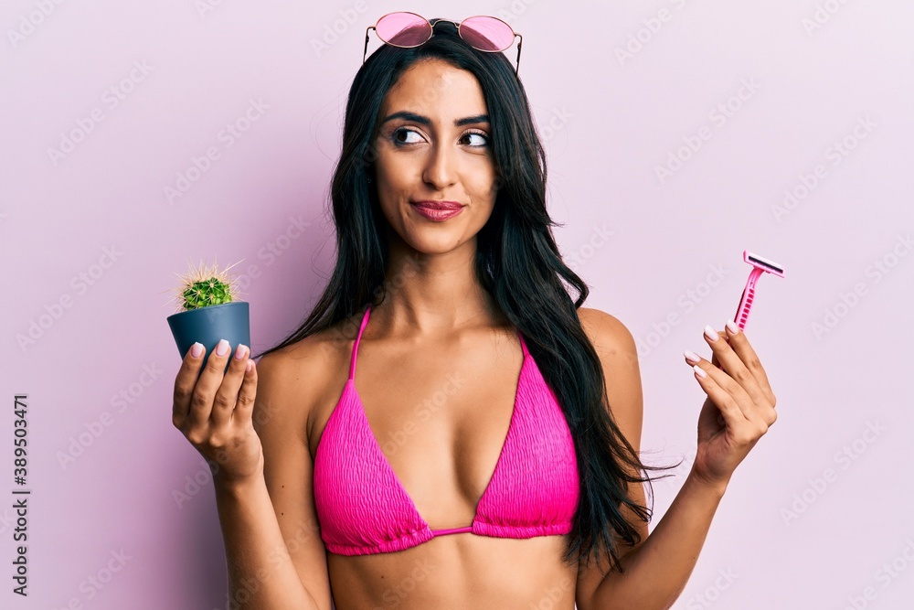 Beautiful hispanic woman wearing bikini holding cactus and razor smiling looking to the side and staring away thinking. Beautiful hispanic woman wearing bikini holding cactus and razor smiling looking to the side and staring away thinking.
