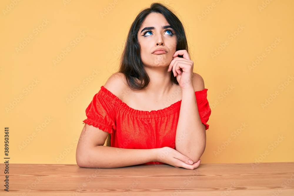 Brunette teenager girl wearing casual clothes sitting on the table serious face thinking about question with hand on chin, thoughtful about confusing idea Brunette teenager girl wearing casual clothes sitting on the table serious face thinking about question with hand on chin, thoughtful about confusing idea