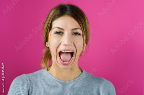 Young adult caucasian woman doing face yoga gymnastics for non-surgical rejuvenation and self-care mouth open with pink background - anti aging concept