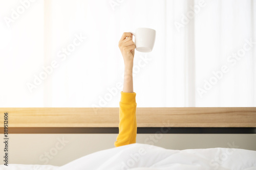 Woman waking up in the morning on bed. She hiding under the blanket and stretching out arms with holding a cup of coffee