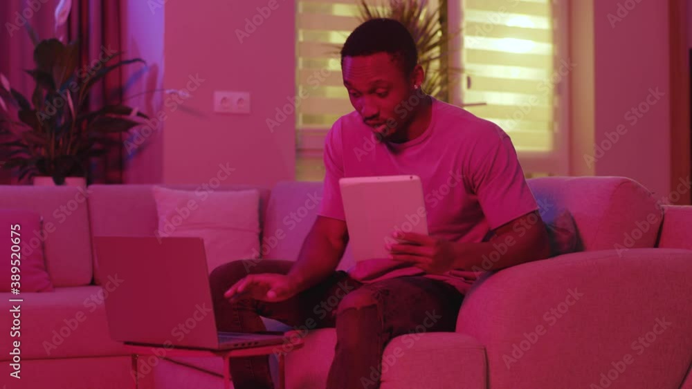 Multitasking. Neon lights. Busy afro-american freelancer doing multiple distant jobs simultaneously getting angry coping with several tasks. Home interior. Remote communication. Multitasking. Neon lights. Busy afro-american freelancer doing multiple distant jobs simultaneously getting angry coping with several tasks. Home interior. Remote communication.