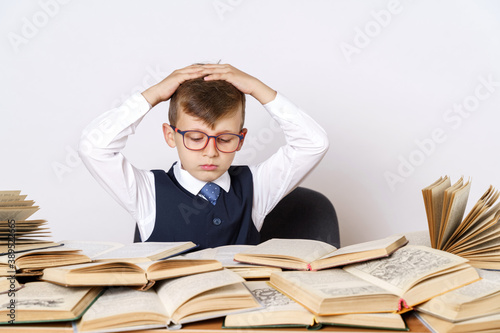 The student does his homework, holds his head with his hands and looks at the camera. There are many open books on the table.