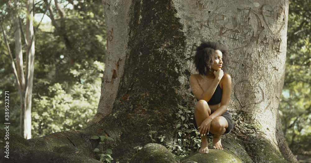 A young girl stoops on the base of a large tree trunk with graffiti written on it