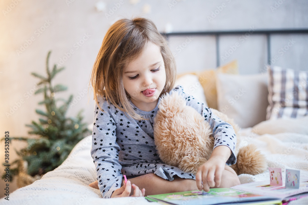 The room is decorated for the new year, a girl is sitting on the bed with a bear, a plush toy is playing school, reading a book, leafing through, a preschooler The room is decorated for the new year, a girl is sitting on the bed with a bear, a plush toy is playing school, reading a book, leafing through, a preschooler