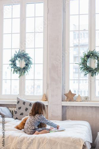 The room is decorated for the new year, a girl is sitting on the bed with a bear, a plush toy is playing school, reading a book, leafing through, a preschooler