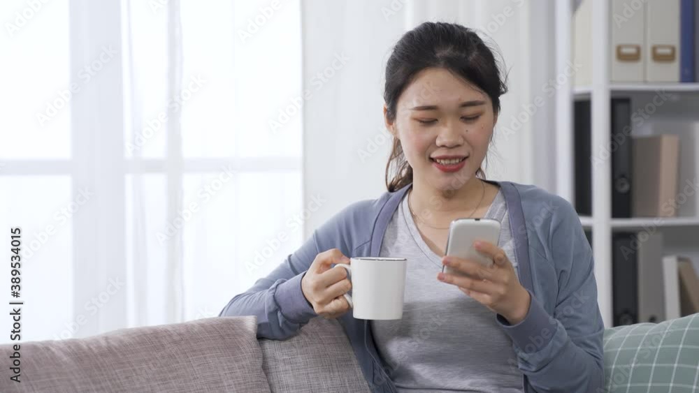 elegant asian young girl sitting on sofa with a cup of hot drink is watching funny video on mobile phone. elegant asian young girl sitting on sofa with a cup of hot drink is watching funny video on mobile phone.